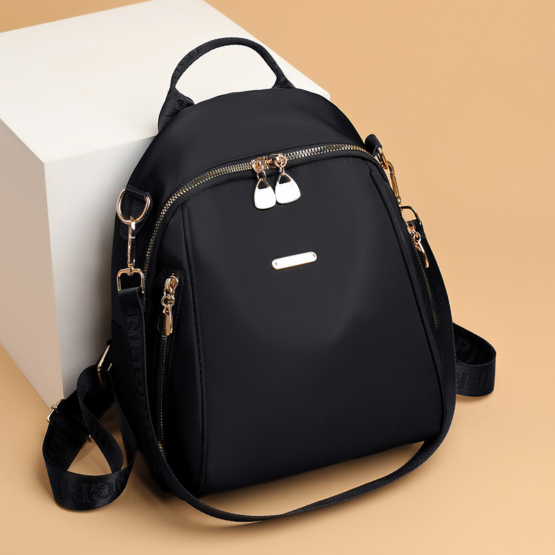 Elegant black SKUGG women’s mini backpack handbag with gold zippers and adjustable shoulder strap, displayed on a beige studio background.